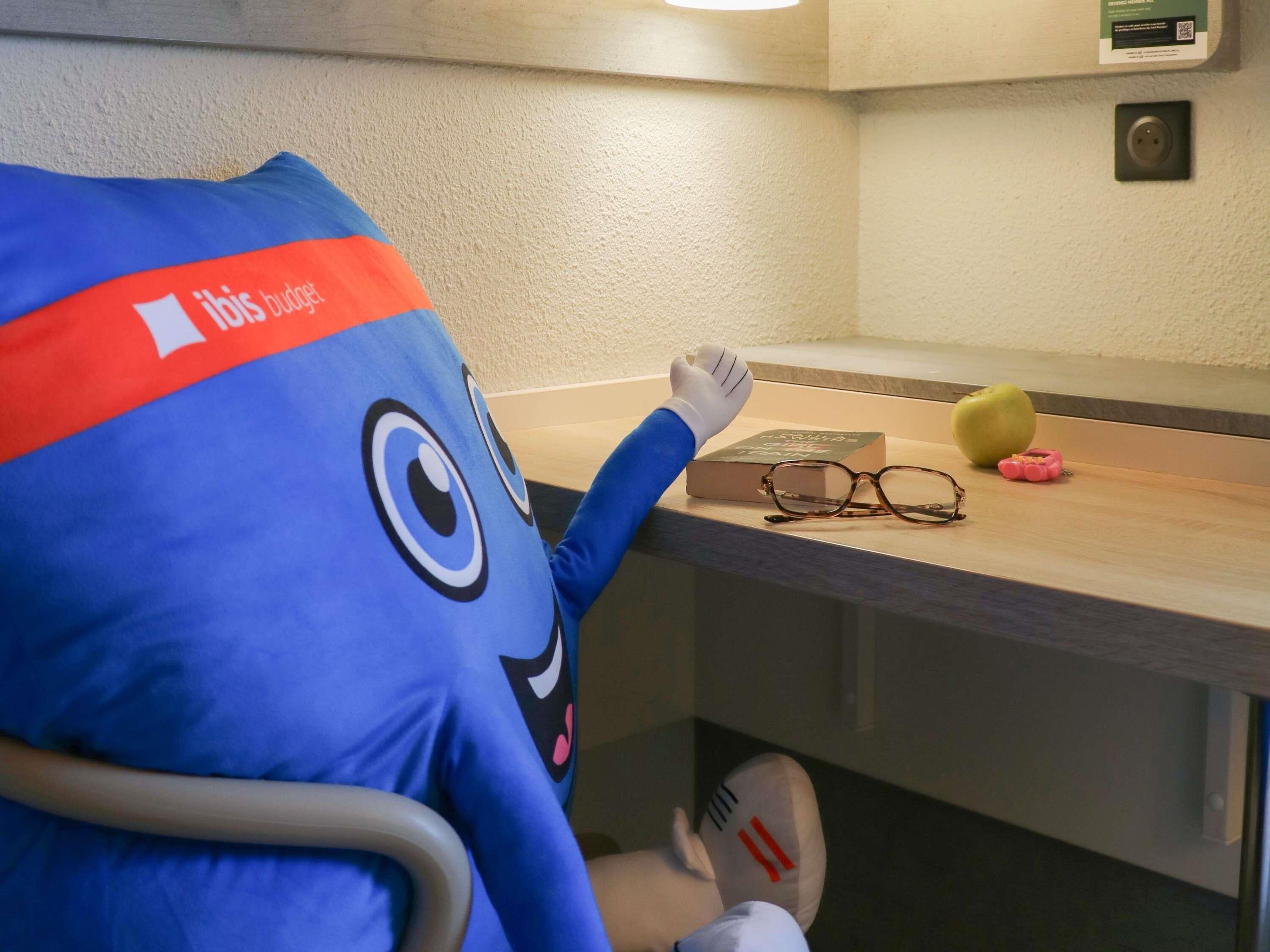 Um personagem mascote azul com o logo vermelho 'ibis budget', um livro, óculos e um carrinho de brinquedo sobre uma superfície de madeira.