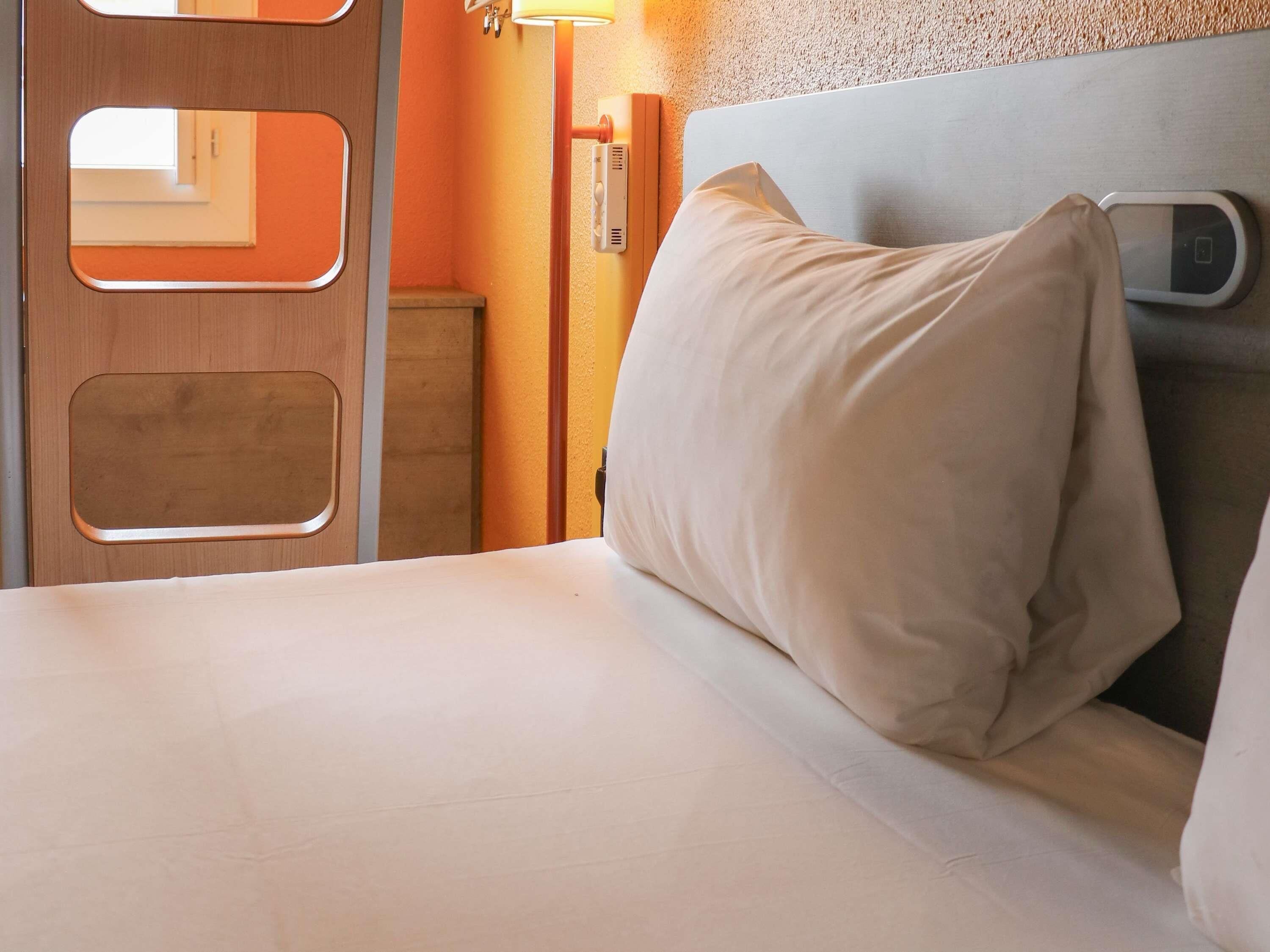 A hotel room with a bed, a nightstand, a wall-mounted control panel, and a door with a mirror.