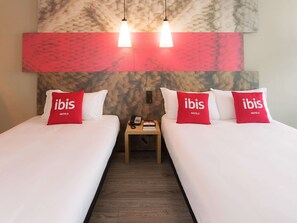 Une chambre d’hôtel avec deux lits, chacun agrémenté d’un oreiller rouge portant l’inscription « ibis HOTELS ». Il y a deux tables de chevet avec des lampes, et un téléphone sur l’une d’elles. La chambre a un plancher en bois et un mur décoré d’un motif marbré au-dessus des lits.