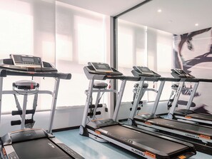 Une rangée de vélos d’appartement dans une salle de sport, avec une grande fenêtre et un mur recouvert de miroirs.