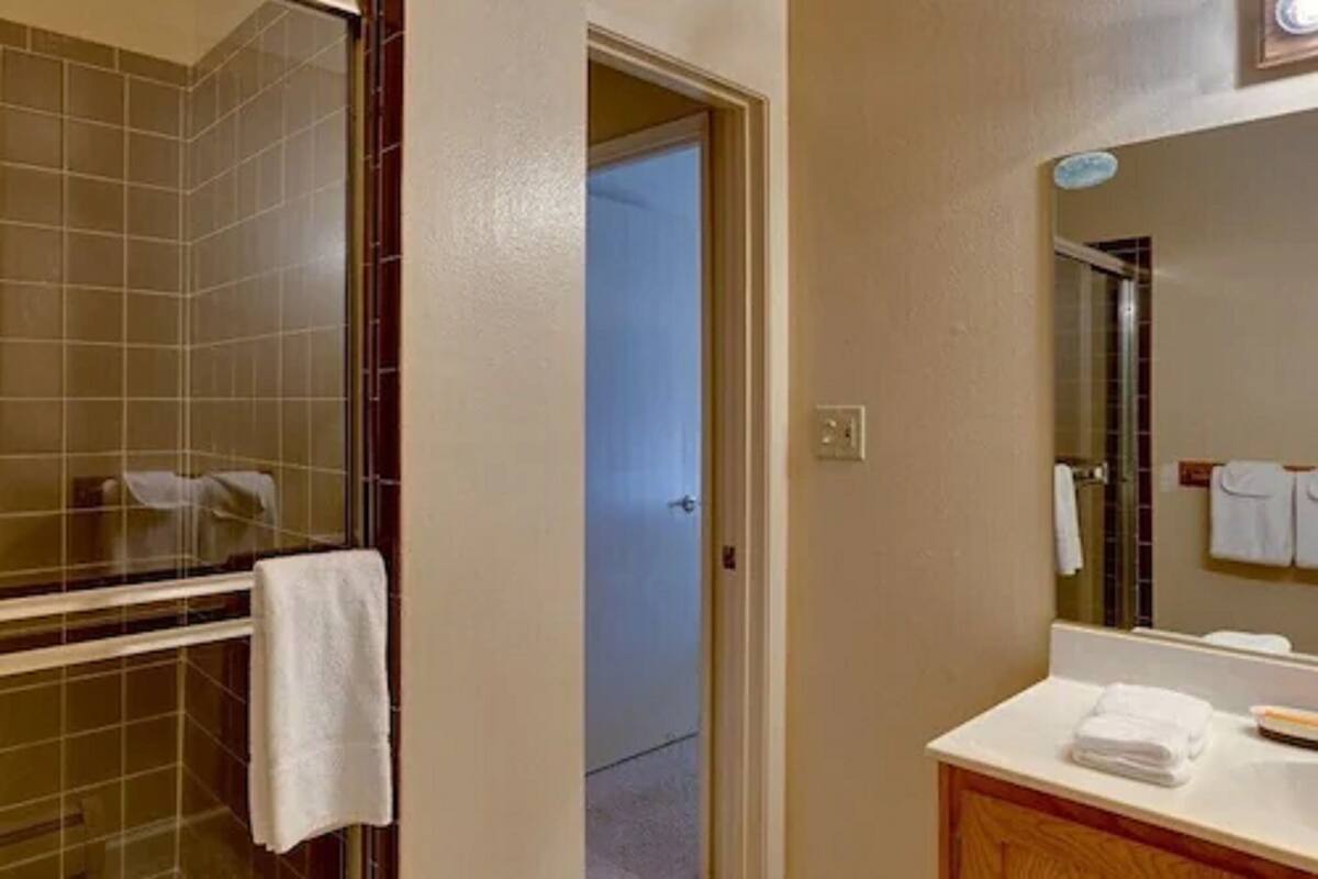 Un baño con zona de ducha azulejada, un lavamanos con espejo y una puerta que da a otra habitación.