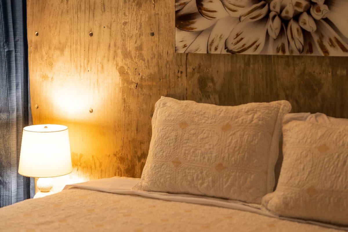 A bedroom with a wooden wall, a floral painting, a bedside lamp, and a bed with pillows.