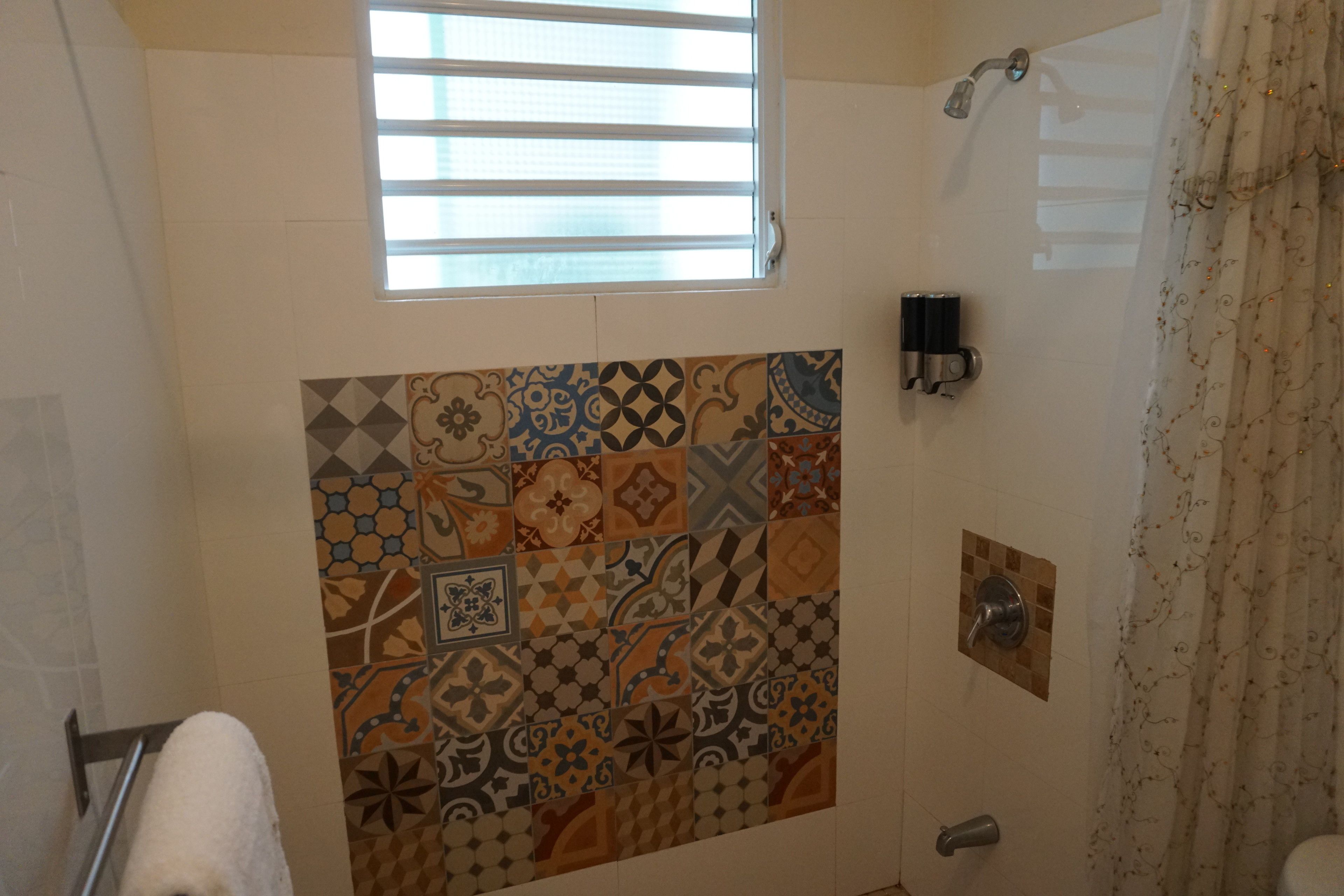 Une salle de bain avec un mur recouvert de carreaux à motifs, une fenêtre avec des stores, une douche et un porte-serviettes.