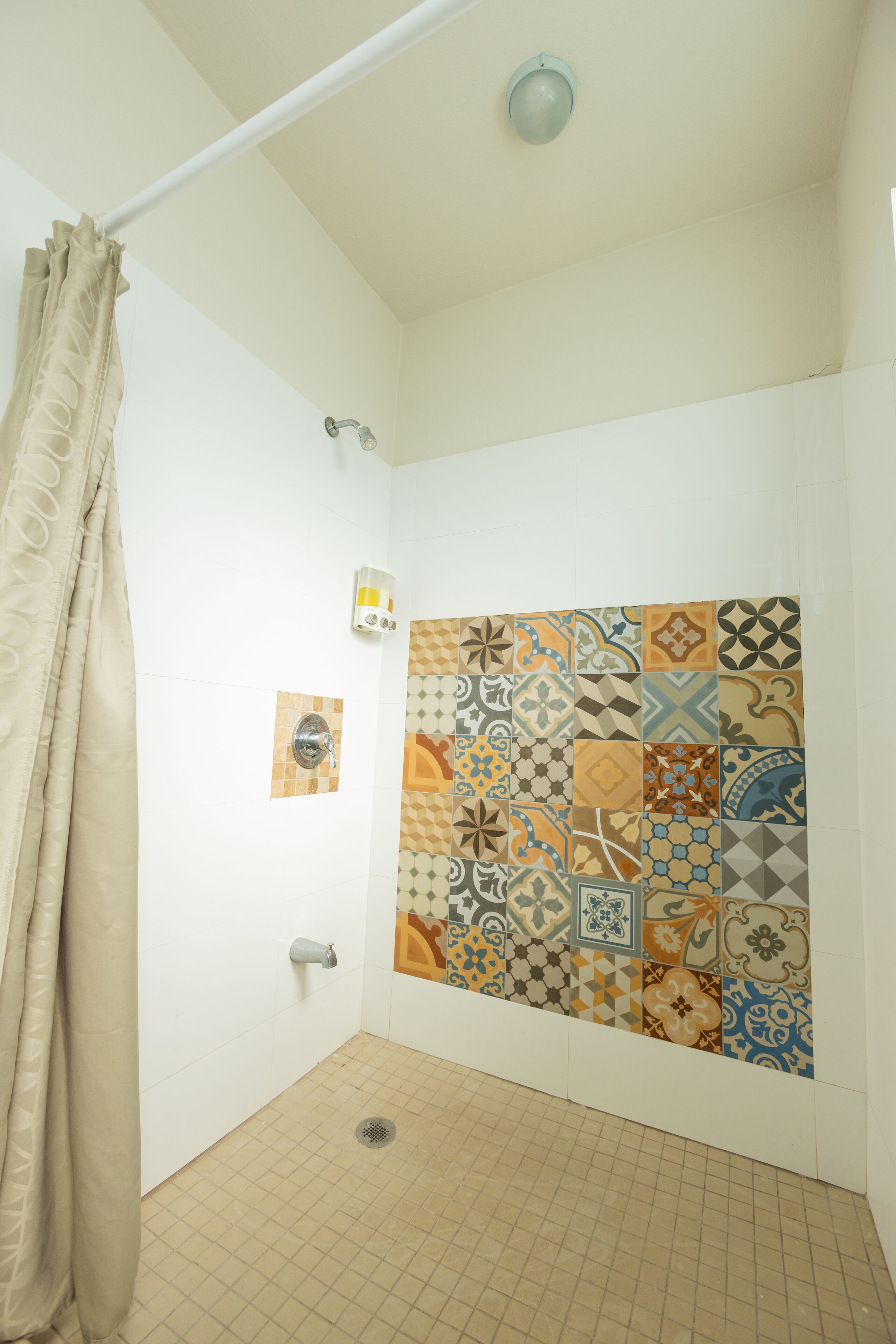 Een douche met een beige gordijn, een in de muur gemonteerde lamp en een betegelde douchewand.