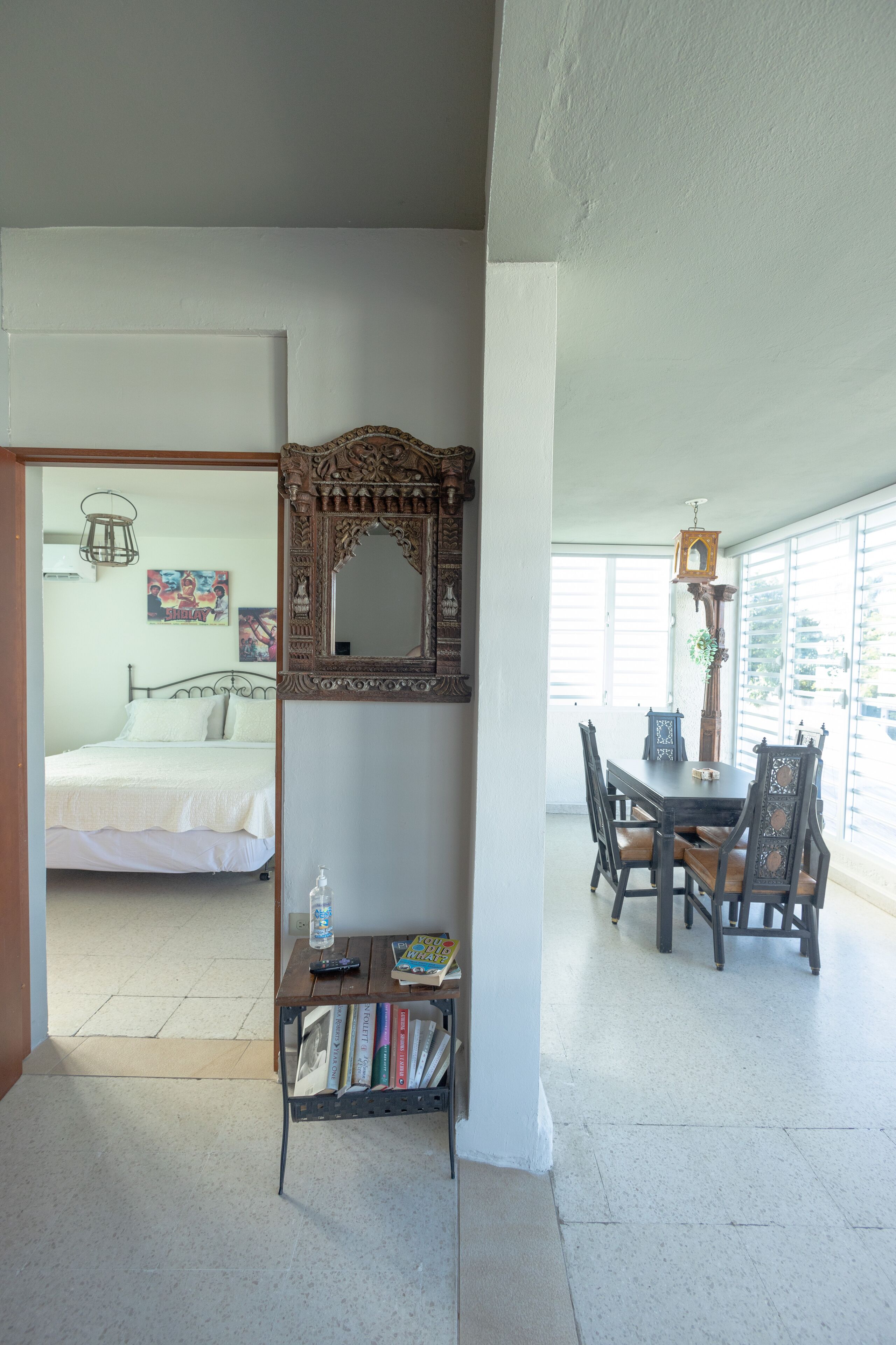 A room with a bed, a dining table with chairs, a bookshelf with books, and a decorative mirror.