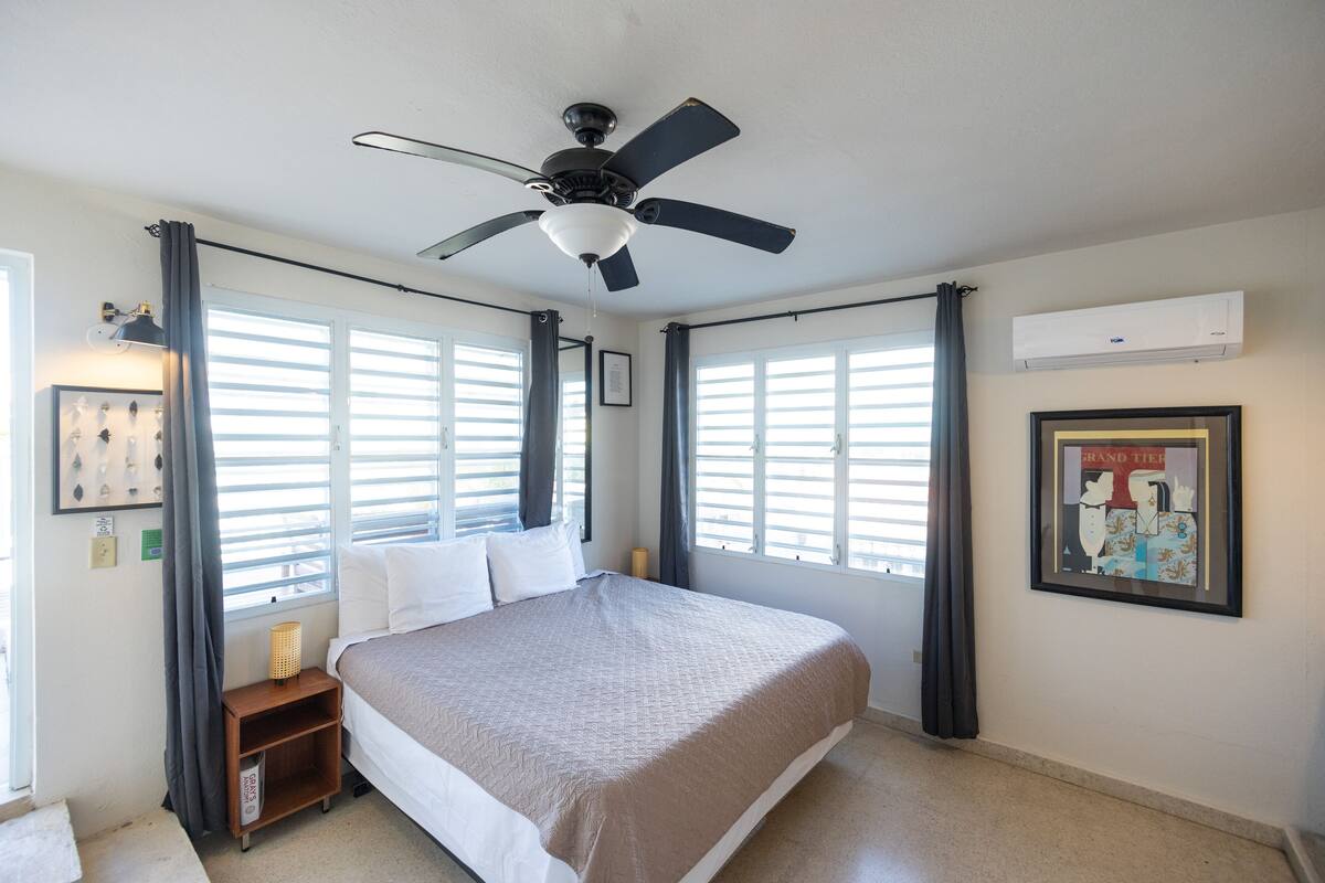A bedroom with a bed, a ceiling fan, a window with blinds, and an air conditioning unit.
