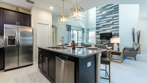 A modern kitchen with a stainless steel refrigerator, a dishwasher, and a granite countertop. There is a dining area with a sofa and a TV. - Solterra - 5431 Solterra Circle (Davenport)