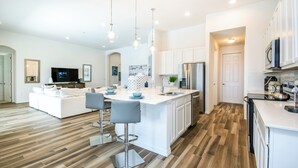 Une cuisine moderne avec des meubles blancs, un îlot central doté de tabourets de bar, des appareils en acier inoxydable et un téléviseur à écran plat fixé au-dessus de l’îlot.