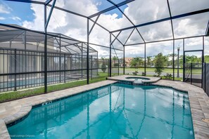 A swimming pool with a glass enclosure, surrounded by a paved deck and a covered area with a roof.