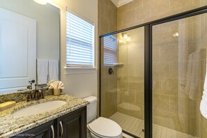 Une salle de bain dotée d’un lavabo avec un plan de toilette en granit, de toilettes, d’une douche à parois de verre et d’une fenêtre avec des stores.