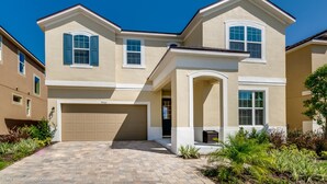 A two-story house with a beige exterior, a garage, and a well-maintained front yard with various plants. - Solara Resort - 9063 Sunshine Ridge Loop (Kissimmee)