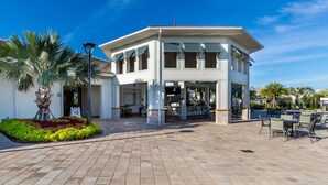 A covered outdoor seating area with tables and chairs, a palm tree, and a building with a white facade and multiple windows.