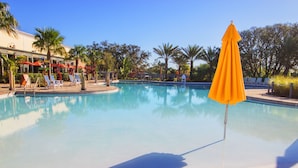 A clear swimming pool with lounge chairs and umbrellas, surrounded by palm trees and a building with outdoor seating.