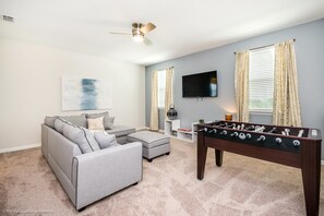Un salon avec un canapé gris, un pouf et une table de baby-foot.
