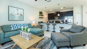 A modern living room with a sofa, ottoman, and coffee table, a kitchen with dark cabinets and stainless steel appliances, and a ceiling fan. - Storey Lake - 3175 Pequod Place (Kissimmee)