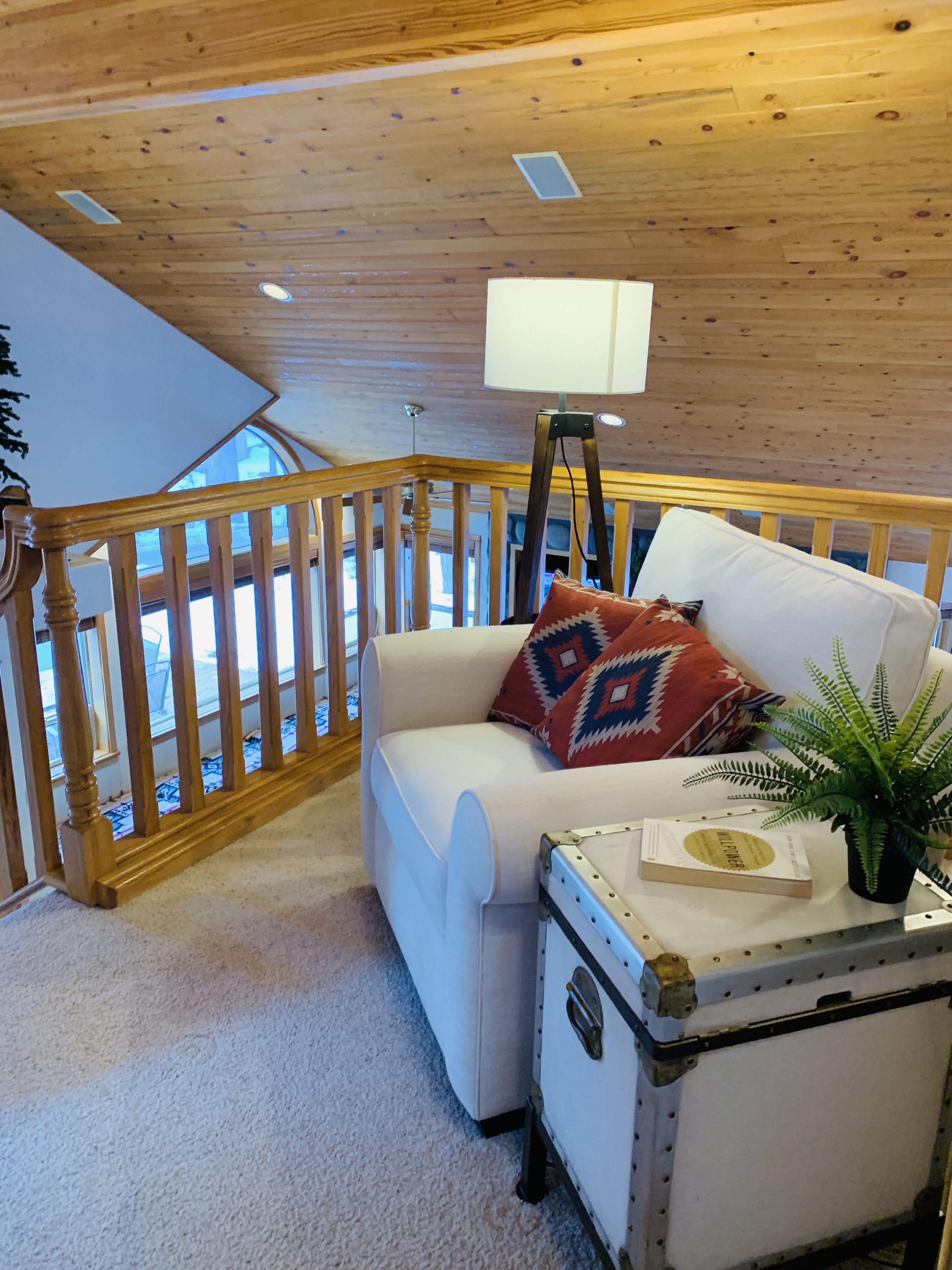 Ein gemütliches Zimmer mit Holzbalkendecke, einem weißen Sofa mit gemustertem Kissen, einem kleinen Tisch mit Buch und Pflanze sowie einer Holzwandleiste.