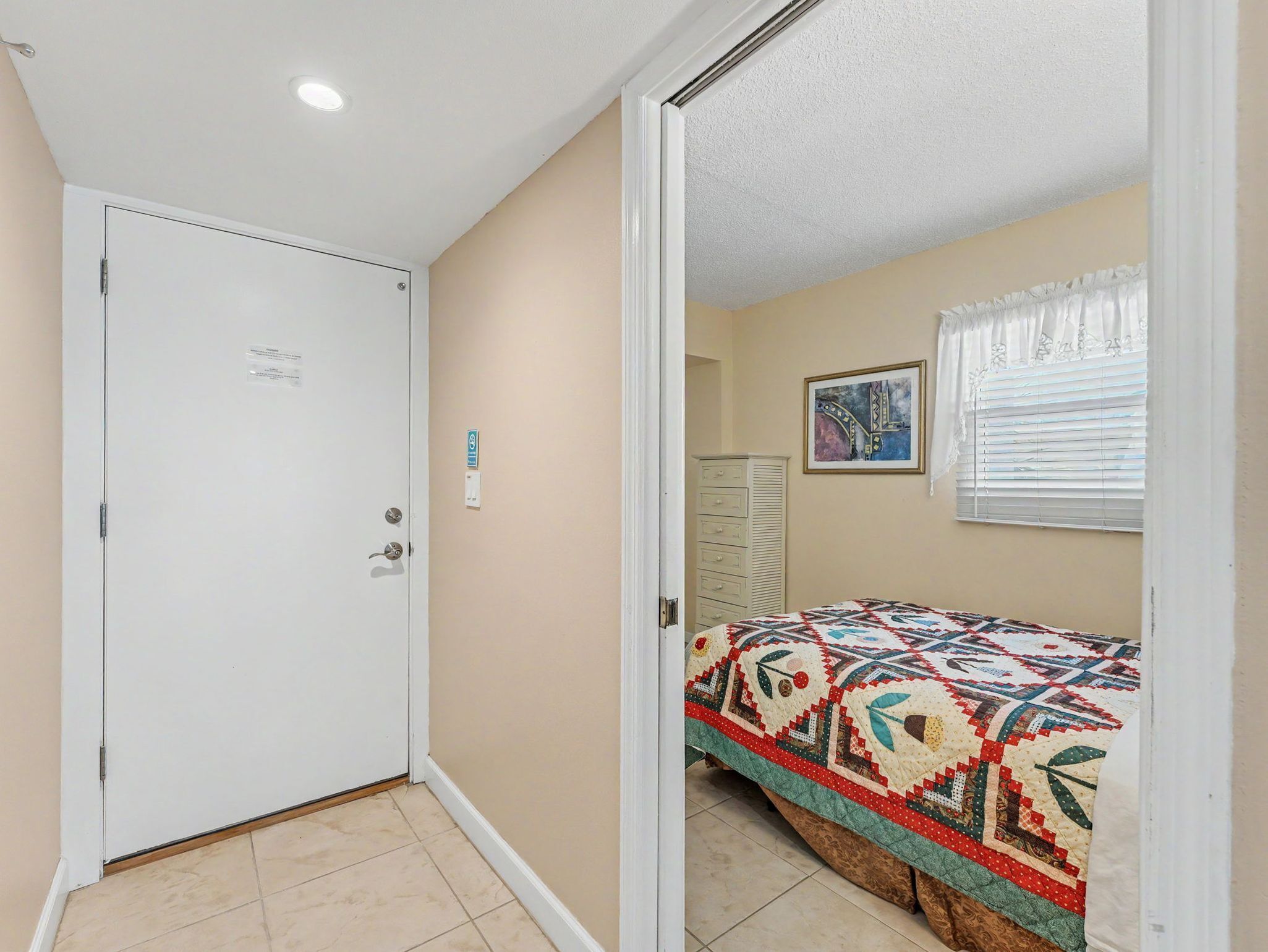 A bedroom with a bed, a door, a window with blinds, and a painting on the wall.