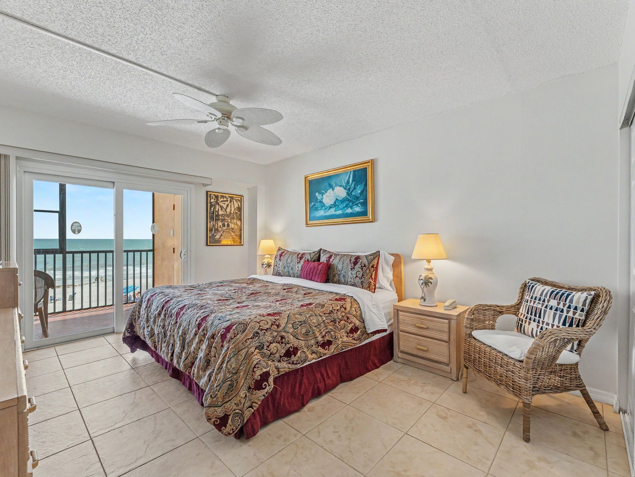 A bedroom with a bed, two bedside tables with lamps, a wicker chair, a ceiling fan, and a door leading to a balcony with a view of the beach.