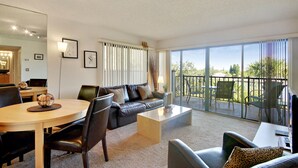 A modern living room with a sofa, coffee table, and dining area. It features large windows and a balcony with outdoor furniture.