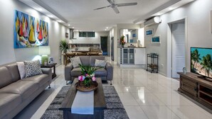 A modern living room with a sofa, coffee table, and a TV. There is a kitchen in the background with a dining area.