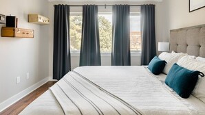 Un dormitorio con una cama grande, cortinas oscuras, cabecera y una ventana con vistas a árboles y edificios.