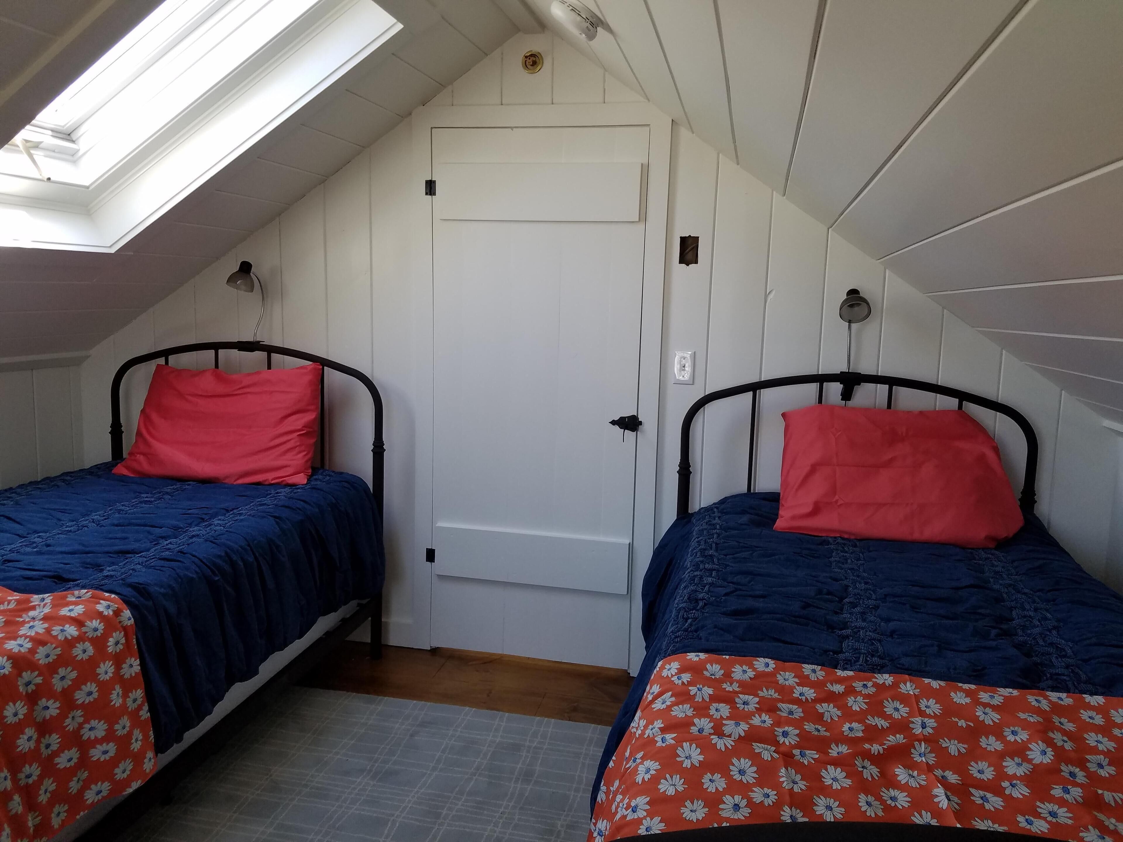 Un alojamiento pequeño, con una sola habitación y dos camas, cada una con almohada roja y ropa de cama azul. Hay un tragaluz sobre una cama y una puerta blanca en el centro.