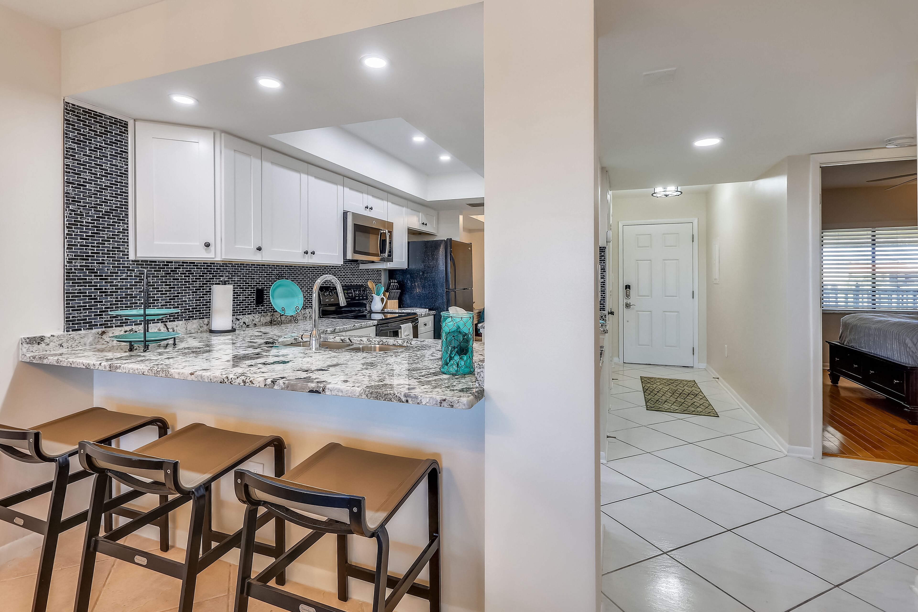 A modern kitchen with white cabinets, a black and white backsplash, and a marble countertop. There are three bar stools and a refrigerator.