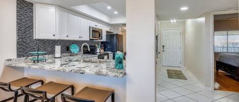 A modern kitchen with white cabinets, a black and white backsplash, and a marble countertop. There are three bar stools and a refrigerator.