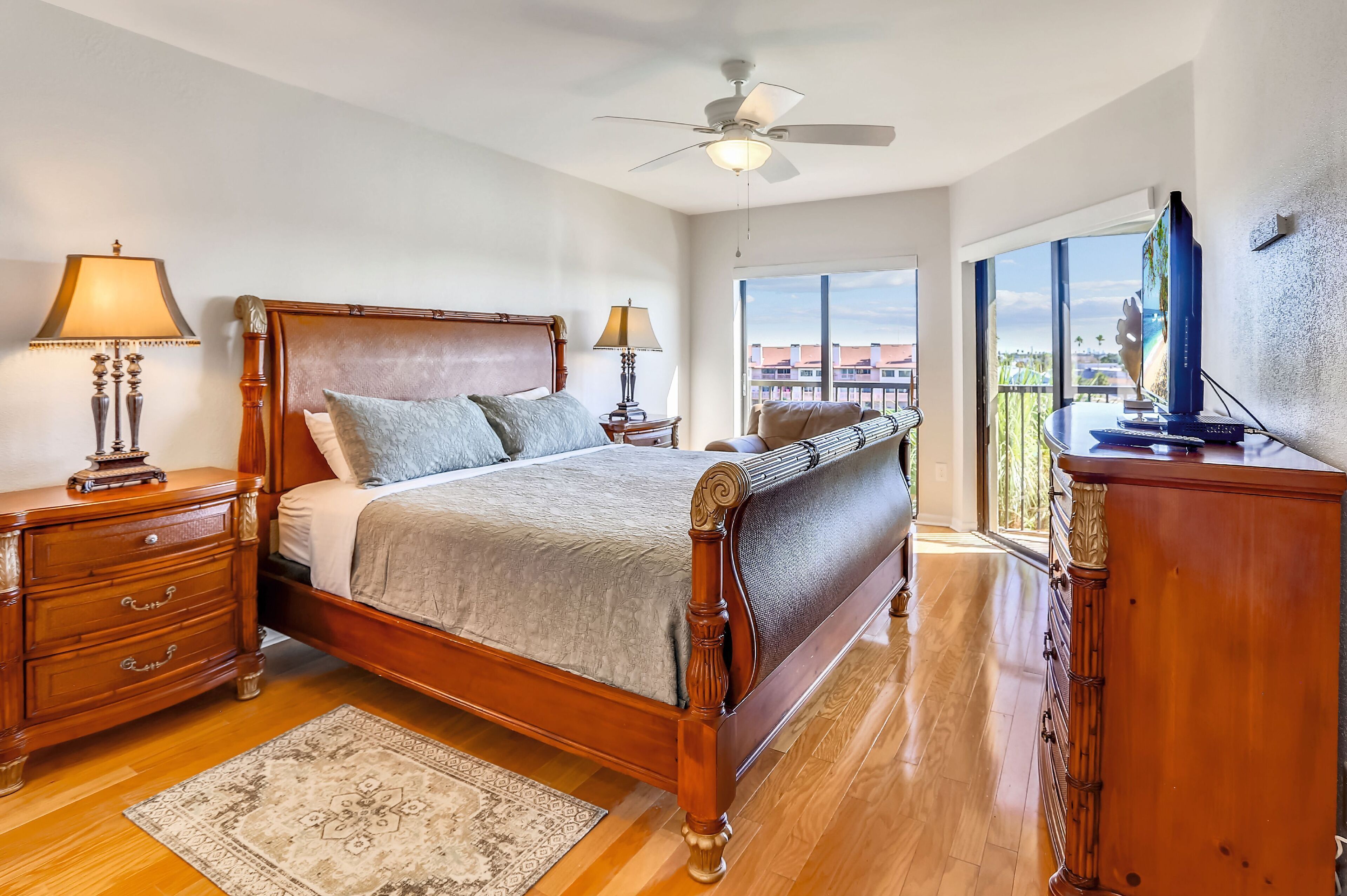 A spacious bedroom with a large bed, wooden headboard, two bedside tables with lamps, a dresser, and a television on a stand.
