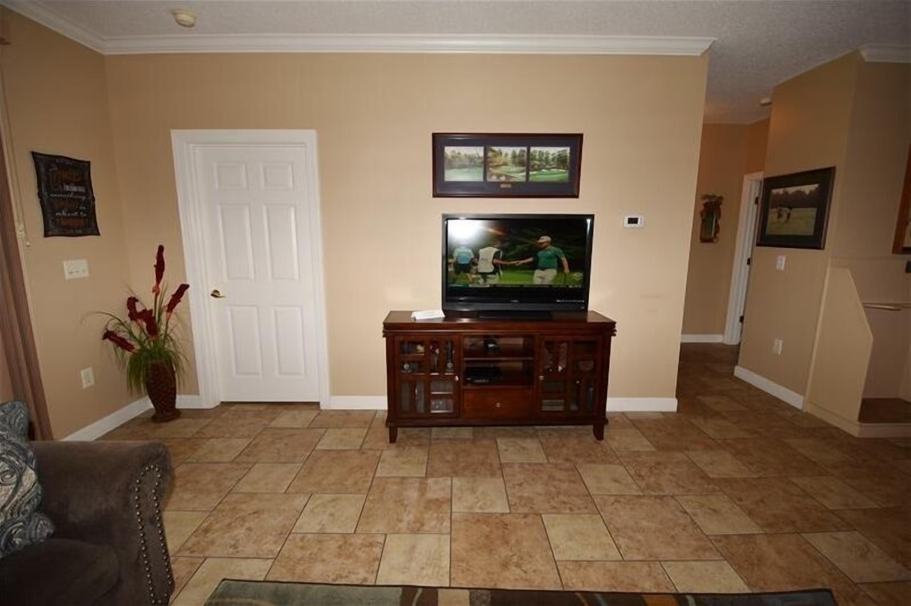 A living room with a television on a wooden stand, beige walls, a door, and a potted plant.