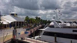 Un puerto deportivo con un yate blanco amarrado, un edificio cubierto con techo inclinado y palmeras al fondo.
