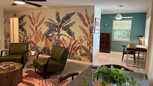 Una sala de estar con un mural de hojas tropicales, dos sillones verdes, una mesa de centro y un comedor con ventanal.