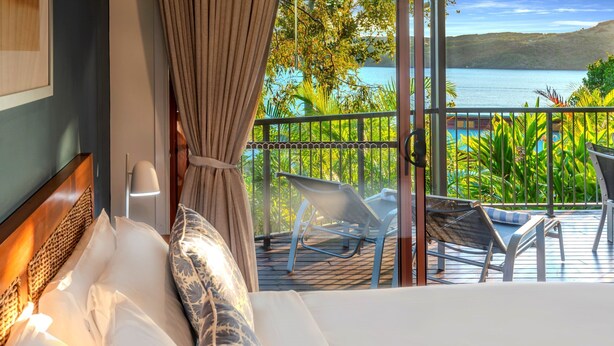 Een hotelkamer met een bed, uitzicht op een balkon met stoelen en een zwembad, en een prachtig uitzicht over een meer en heuvels.