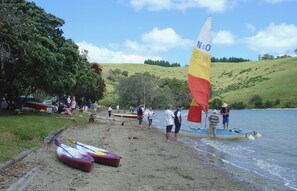 A lakeside setting with canoes and a sailboat, people near the water, and a grassy hill in the background. - The Yellow Bach 90m to Campbells Beach; kayaks, wifi, Sky, Netflix, sleeps 4 (Campbells Beach)