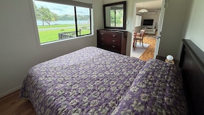 A bedroom with a large bed, a dresser, a mirror, and a view of a park and lake through a window. - The Yellow Bach 90m to Campbells Beach; kayaks, wifi, Sky, Netflix, sleeps 4 (Campbells Beach)