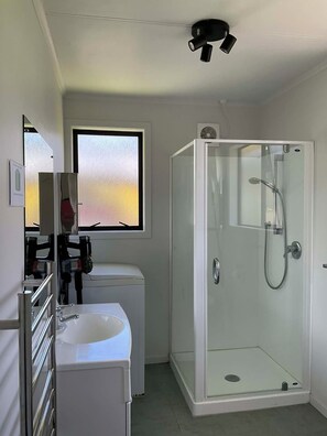 A bathroom with a glass-enclosed shower, a sink, a mirror, and a window with a view. - The Yellow Bach 90m to Campbells Beach; kayaks, wifi, Sky, Netflix, sleeps 4 (Campbells Beach)