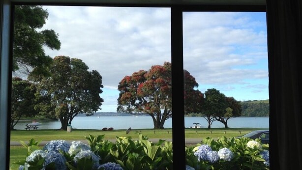 A view through a window of a park with trees, a lake, and a picnic table. - The Yellow Bach 90m to Campbells Beach; kayaks, wifi, Sky, Netflix, sleeps 4 (Campbells Beach)