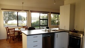 A modern kitchen with a dishwasher, stove, and large windows offering views of trees and a body of water. - The Yellow Bach 90m to Campbells Beach; kayaks, wifi, Sky, Netflix, sleeps 4 (Campbells Beach)