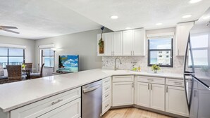 Una cocina moderna con armarios blancos, lavavajillas y un lavabo. Hay una zona de comedor con sillas de mimbre y una mesa, y una ventana con vistas a edificios.
