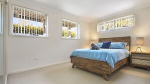 Une chambre à coucher avec un lit en bois, deux fenêtres avec des stores blancs, une table de chevet avec une lampe et un sol recouvert d’une moquette.