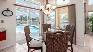 Un comedor con una mesa de vidrio, sillas de mimbre, un candelabro y vista a una piscina.