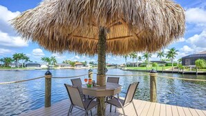 Una terraza de madera con una mesa y sillas bajo un toldo de paja, con vista a un cuerpo de agua, con palmeras y edificios al fondo.