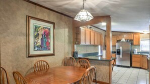 A dining area with a wooden table and chairs, a framed artwork on the wall, and a kitchen with wooden cabinets and a stainless steel refrigerator. - Ooltewah Cabin w/ Grill, Pool Table & Porch! (Ooltewah)