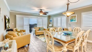 A spacious living room with a large glass dining table, beige sofas, and a ceiling fan.