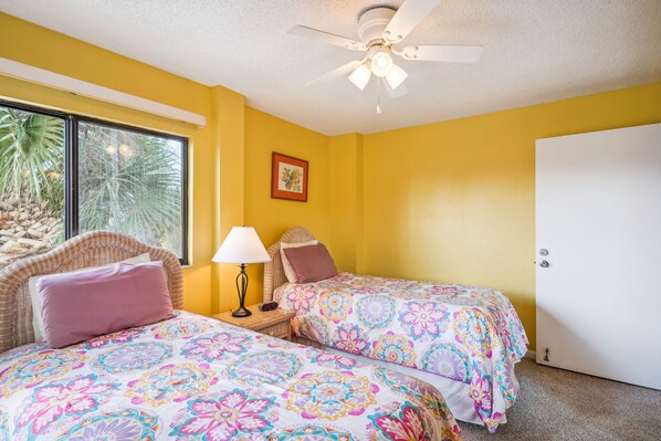 A room with two beds, a ceiling fan, a window with a view of palm trees, and a door. - Land's End 307 building 1 TWO PRIVATE Balconies|Across from POOL|Gulf Views! (Treasure Island)