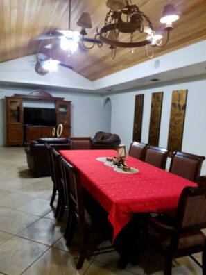 Un coin repas avec une nappe rouge, des chaises en bois, un lustre et un téléviseur en arrière-plan.