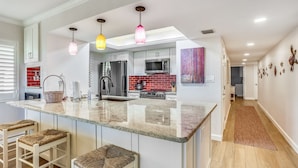 A modern kitchen with a central island, white cabinetry, and a red brick backsplash. There are three bar stools with woven seats, a stainless steel refrigerator, and a microwave above the island.