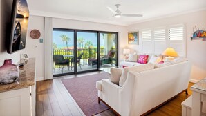 A modern living room with a white sofa, a flat-screen TV, and a large sliding glass door leading to a balcony with outdoor furniture.