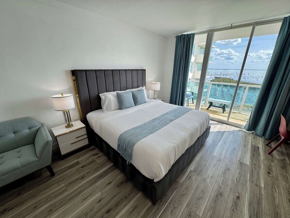 A hotel room with a large bed, a nightstand with a lamp, a chair, and a view of the sea through large windows.