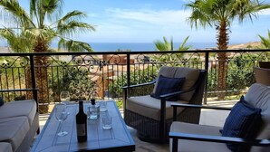 Un balcon agrémenté de meubles en osier, une table avec des verres à vin et une bouteille, et une vue sur la mer et les bâtiments.